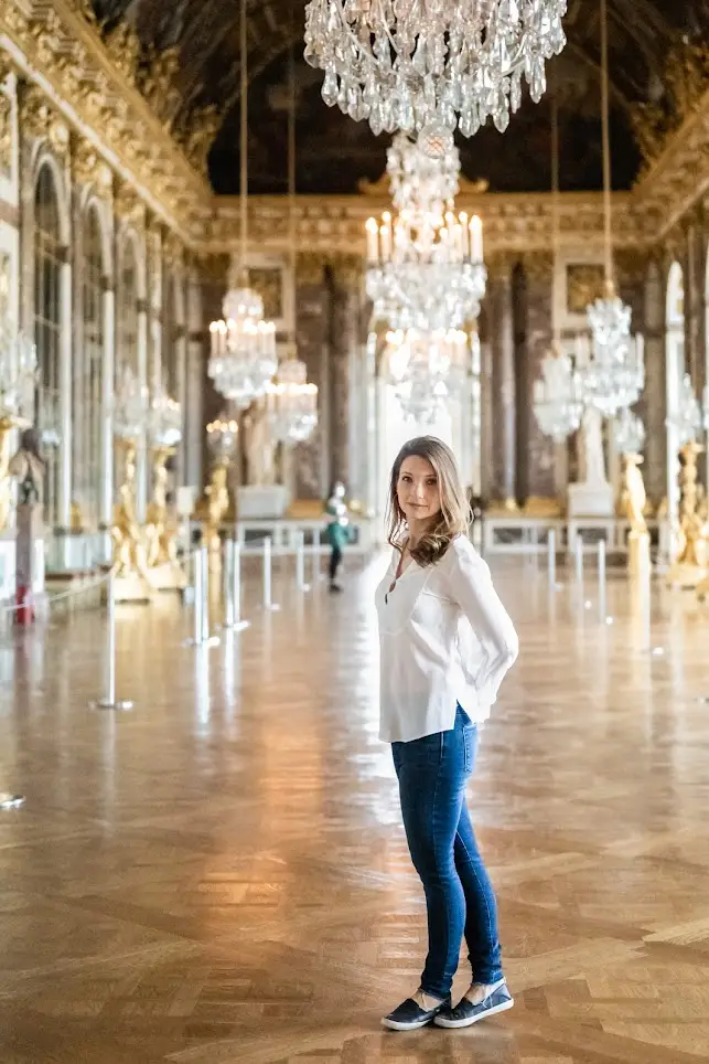 Author standing in the alone in the hall of mirrors. There are big chandeliers and wooden floors. I am wearing jeans and a white shirt.