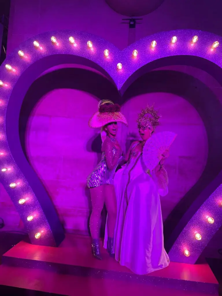 Two guests pose playfully in glittering costumes beneath a glowing heart-shaped frame at the masked ball of Versailles, lit in dramatic pink and purple hues for a theatrical photo moment.