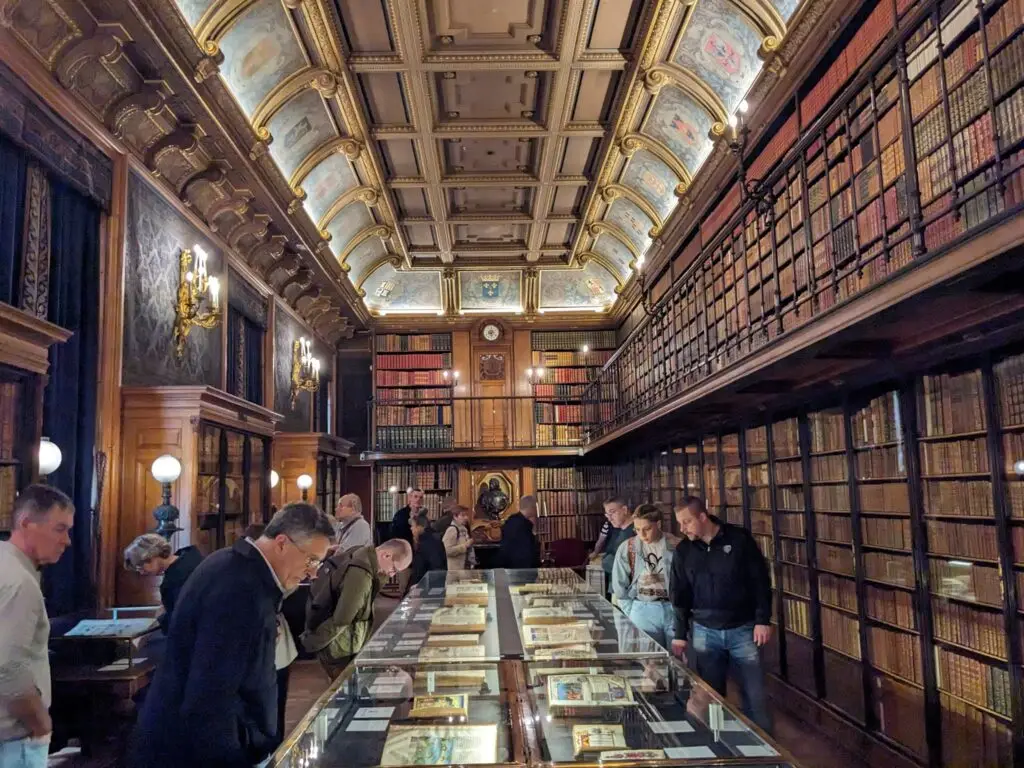 Lavishly decorated library at the Château de Chantilly, featuring floor-to-ceiling bookshelves filled with old volumes, a coffered ceiling with painted panels, and soft, golden lighting. Visitors examine illuminated manuscripts displayed in glass cases, surrounded by dark wood paneling and ornate detailing.