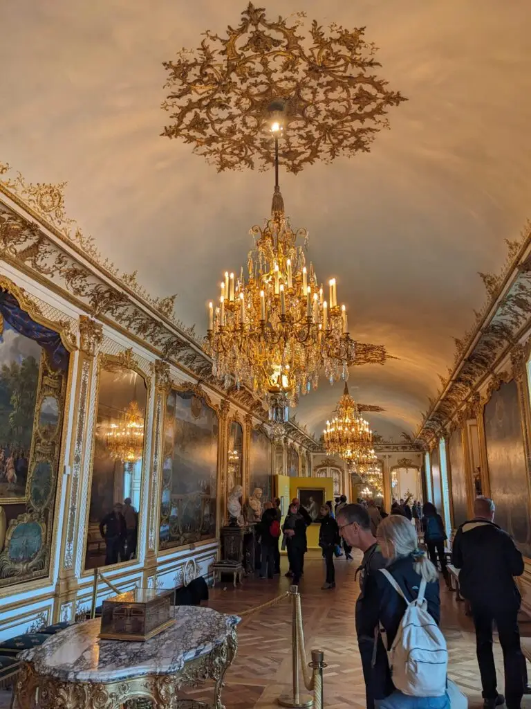Ornate gallery inside the Château de Chantilly, with golden chandeliers, intricate ceiling decorations, and walls lined with massive framed paintings and gilded moldings. Visitors stroll through the opulent hall, taking in the grandeur—despite it being one of the least crowded moments.