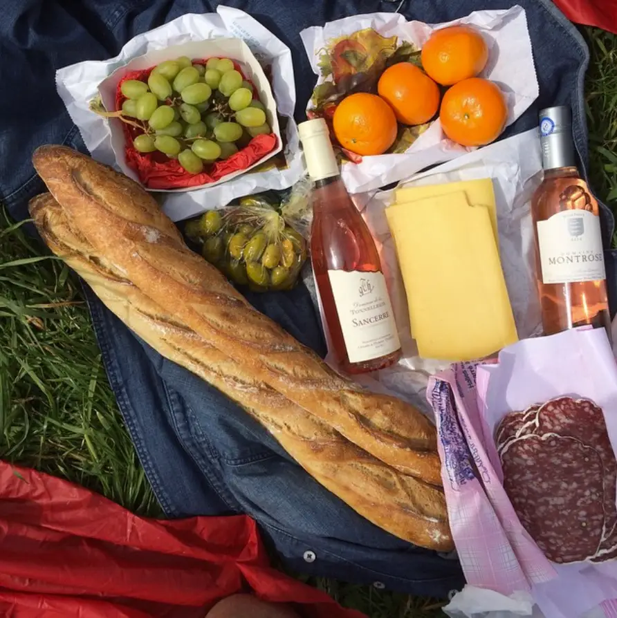 Picnic at Versailles with a spread laid out on a denim jacket in the grass, featuring two baguettes, green grapes, green olives, mandarins, slices of cheese, slices of salami, and two bottles of rosé wine labeled 'Sancerre' and 'Montrose'.