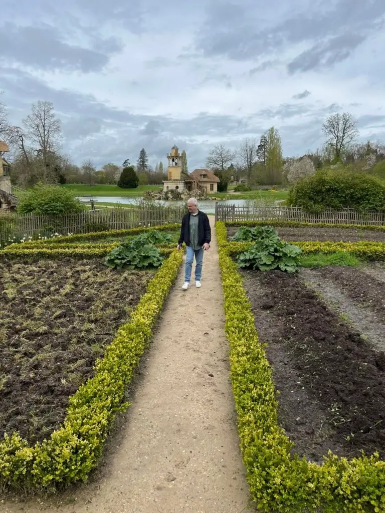 Man walking along a garden path in the Queen’s Hamlet at Versailles, with manicured plots and the Marlborough Tower in the distance.