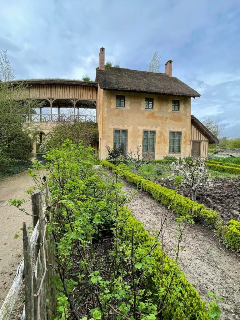 Charming rustic house with a thatched roof and arched walkway in the Queen’s Hamlet at Versailles, surrounded by manicured gardens and young shrubs.