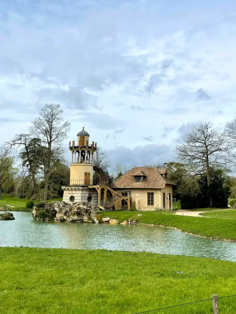 Scenic view of the Marlborough Tower beside a pond in the Queen’s Hamlet at the Palace of Versailles, framed by green lawns and trees. A day at Versailles.