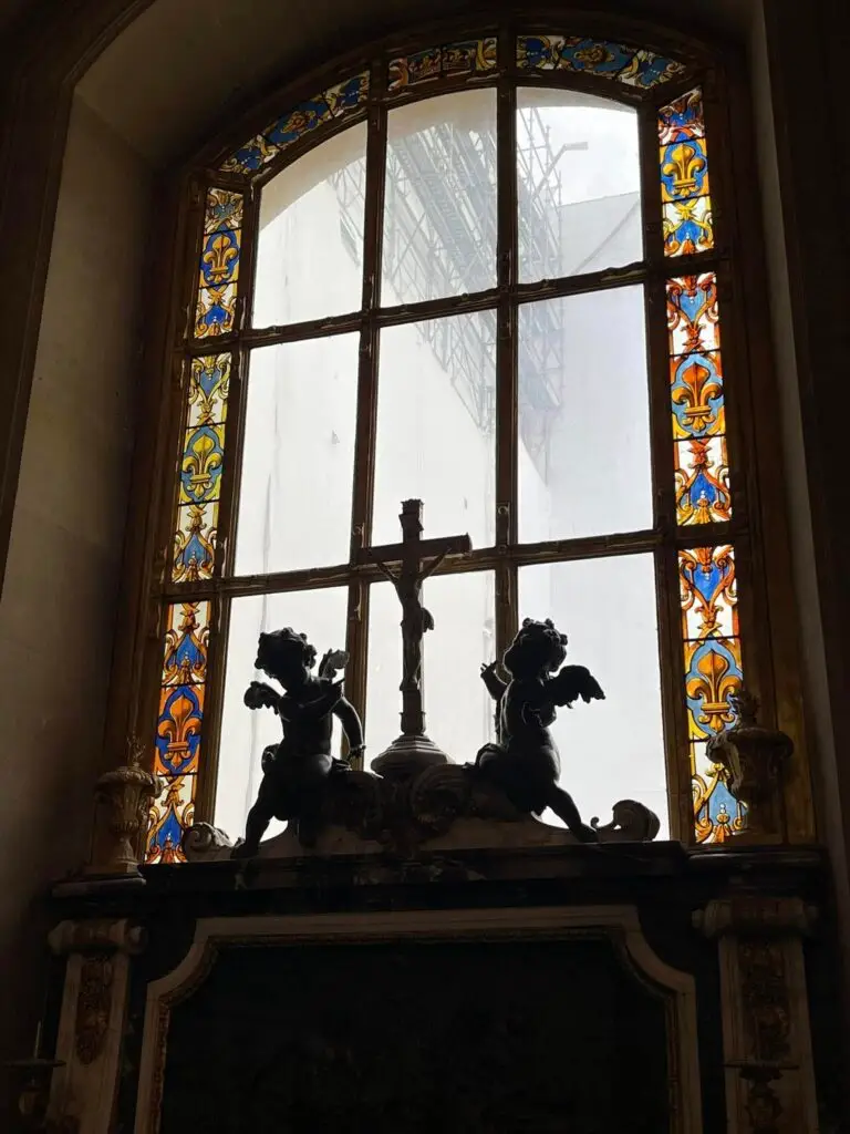 Stained glass window inside the Palace of Versailles framed by fleur-de-lis motifs, with silhouetted cherub statues and a crucifix in the foreground