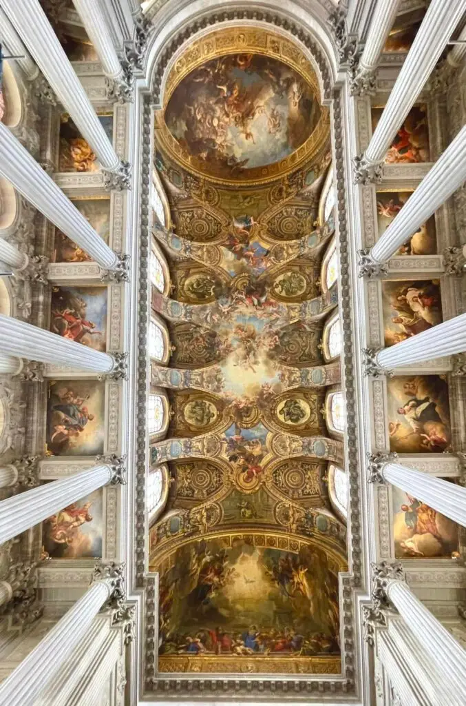 Panoramic ceiling view inside the Royal Chapel at Versailles, displaying intricate Baroque frescoes and detailed gilded frames between white columns.