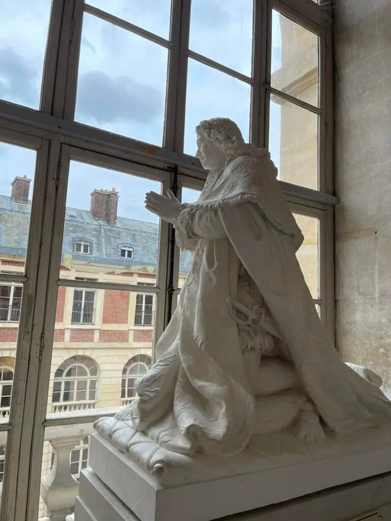 White marble statue of a kneeling man in prayer beside a large window at the Palace of Versailles, with classical buildings visible in the background.