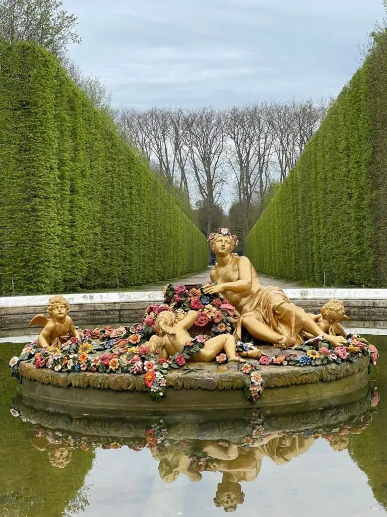 Golden fountain statue of a goddess with cherubs surrounded by colorful flowers, set in a tranquil pool within the Versailles gardens.