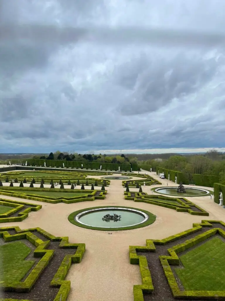 Aerial view of the geometric ornamental gardens at the Palace of Versailles, featuring manicured hedges, fountains, and statues under a cloudy sky