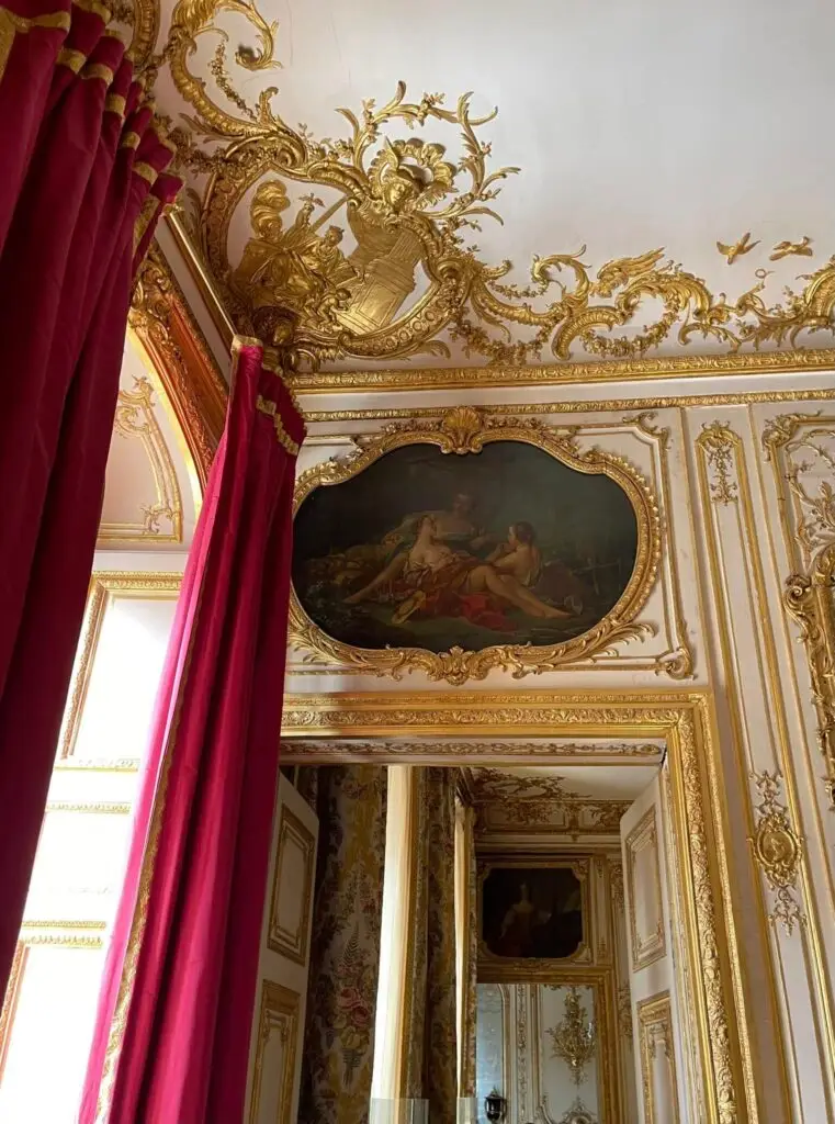 Detail of a Rococo-style interior at the Palace of Versailles with gold gilding, red velvet curtains, and a classical painting above an ornate doorway.
