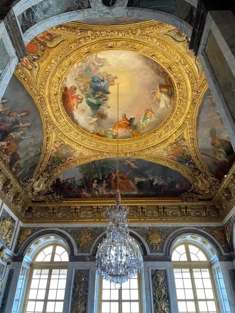 Elaborate ceiling fresco framed by golden ornamentation in the Peace Room at Versailles, with a crystal chandelier and arched windows below.