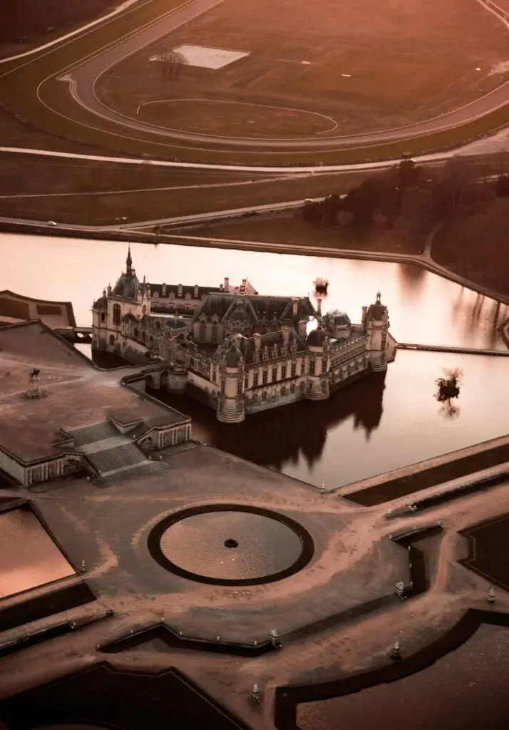 Aerial sunset view of Chantilly Castle and its symmetrical French gardens, surrounded by water and forested parkland.