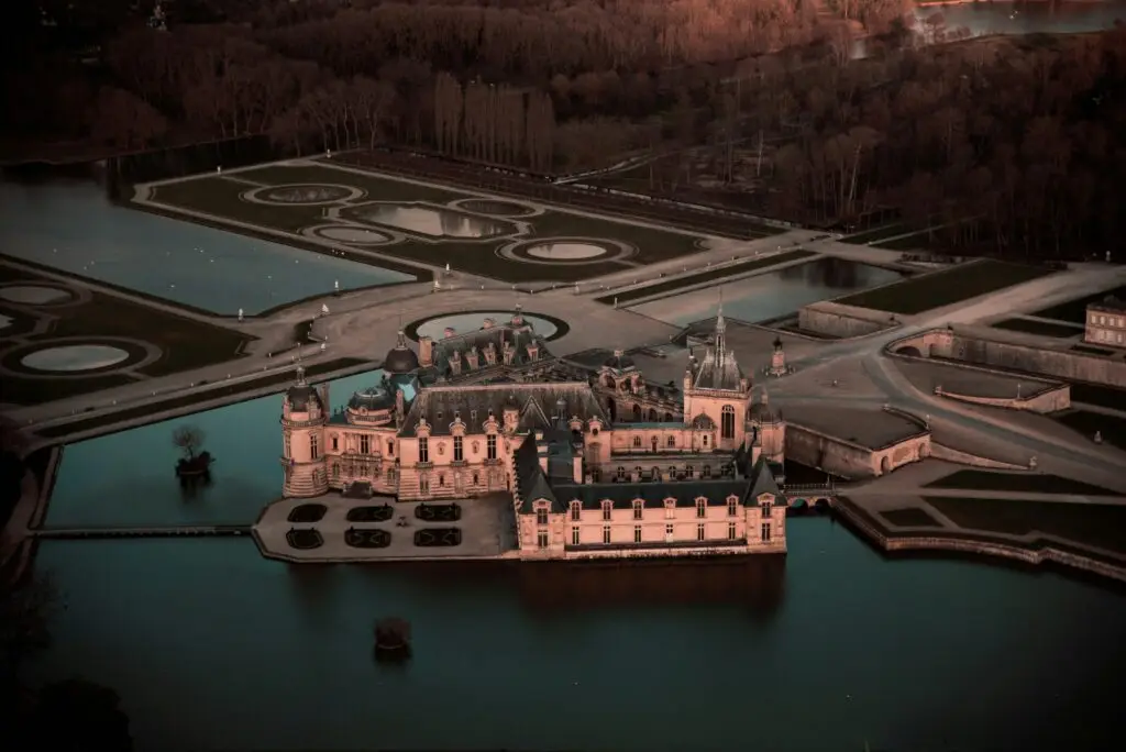 Golden-hour aerial view of Chantilly Castle with reflective moats, circular fountains, and formal garden layout in the Château de Chantilly estate.