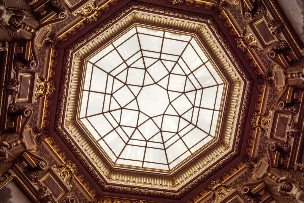 Ornate octagonal glass ceiling inside Chantilly Castle, featuring a geometric pattern and surrounded by lavish Baroque-style gold and bronze molding with sculpted figures.