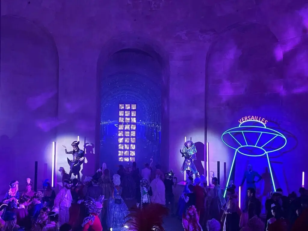 A futuristic theme at the masked ball of Versailles, with neon lights, sci-fi costumes, and a glowing sign reading “Versailles” set against the palace’s stone arches, as guests in baroque and fantasy attire look on.