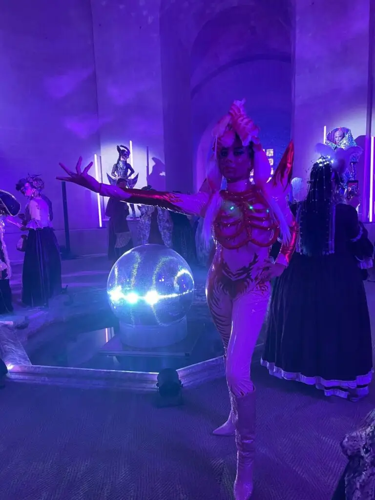 A performer in a futuristic, body-hugging costume strikes a dramatic pose beside a glowing disco ball at the masked ball of Versailles, surrounded by guests in elaborate historical and fantasy attire under purple lighting.