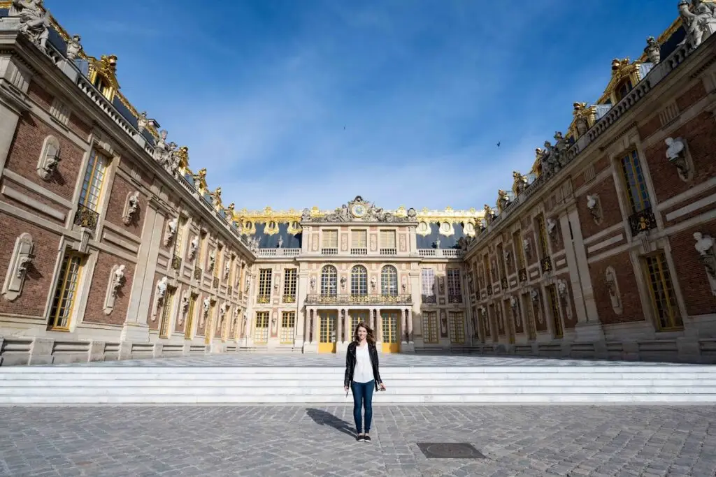 Katie Danger standing in the main courtyard of the Palace of Versailles, France, surrounded by Baroque architecture with golden accents under a bright blue sky.