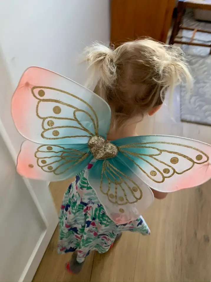 Young child with messy pigtails wearing colorful fairy wings and a floral dress, walking down a hallway on a wooden floor.