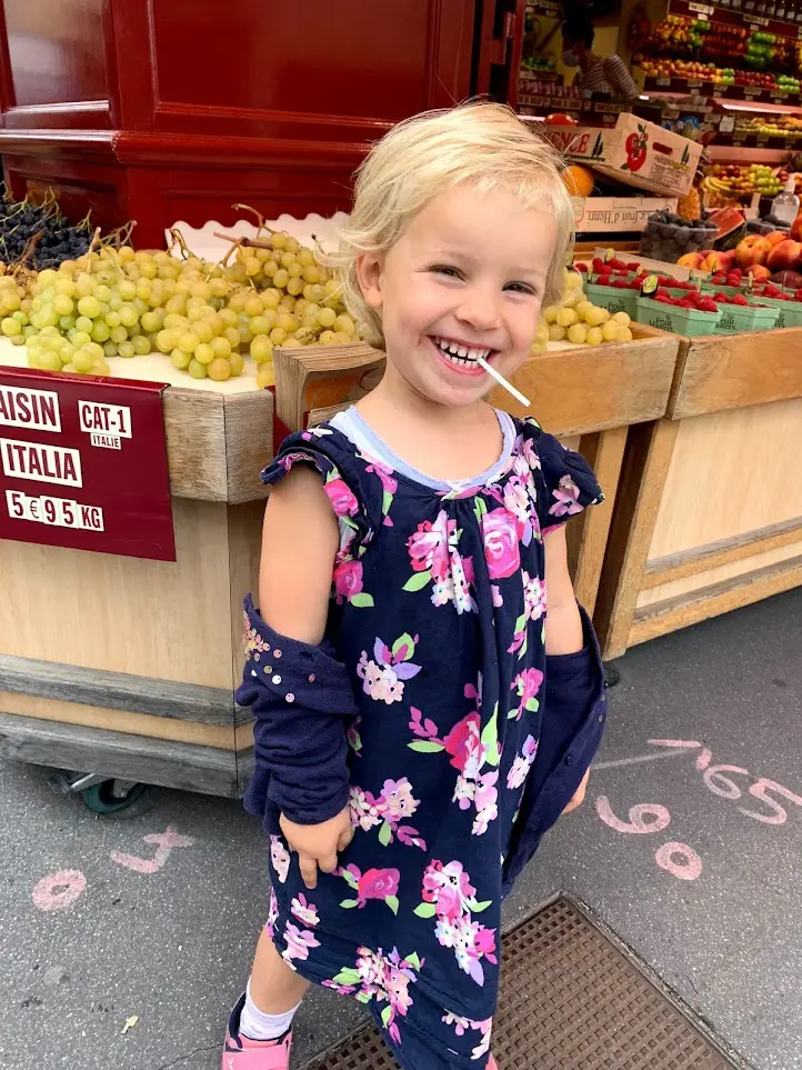 A smiling young girl stands in front of a colorful fruit stand at a local market in Versailles, holding a lollipop. Visiting the markets is one of the most charming family weekend activities near Versailles, offering fresh produce and a taste of everyday French life.