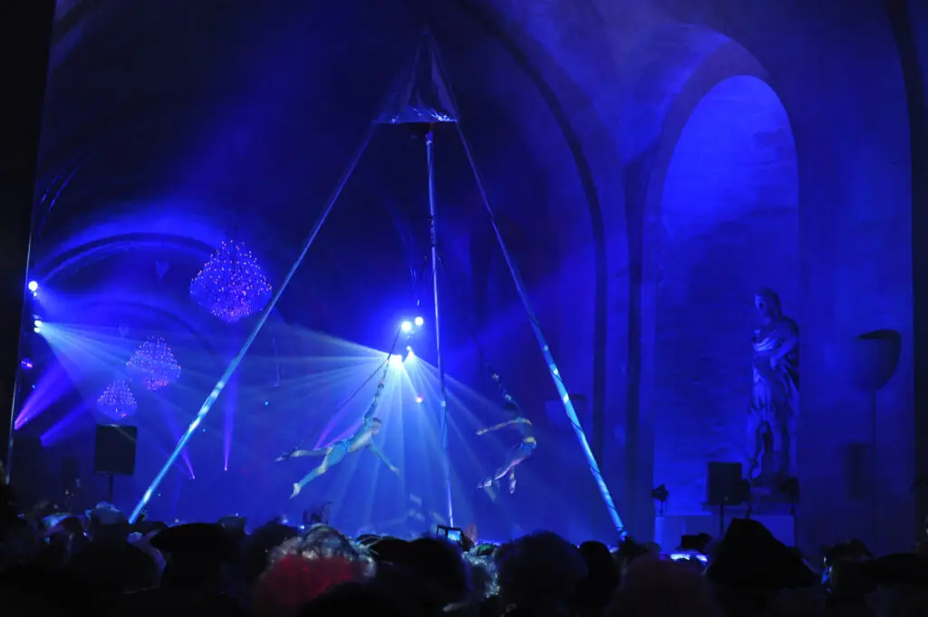 Two aerial acrobats perform high above a costumed crowd at the Versailles masked ball, illuminated by dramatic blue lighting and chandeliers in a grand arched hall with classical statues.