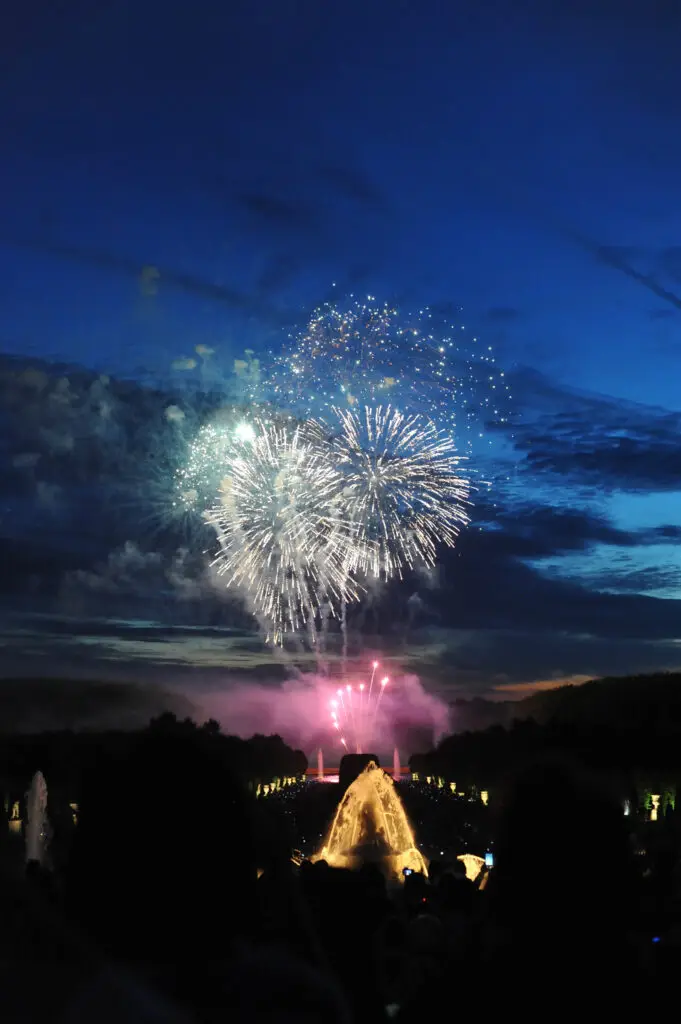 A dazzling firework display lights up the night sky above the Grand Canal during the masked ball of Versailles, as golden fountains shimmer and spectators watch in awe across the palace gardens.