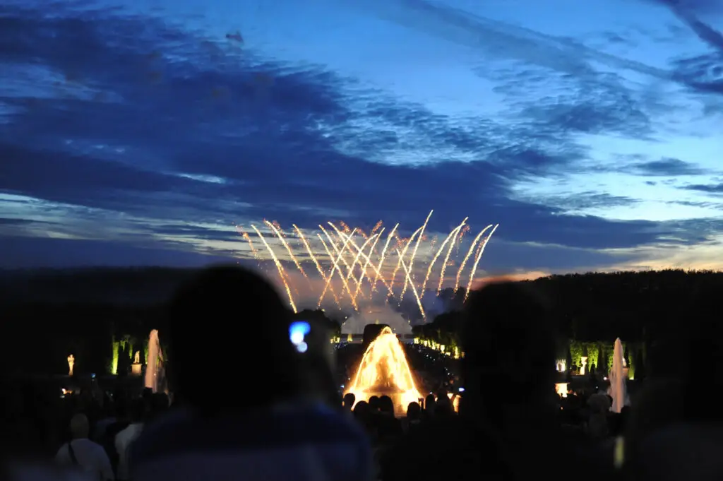 Golden fireworks arc across the evening sky above the Grand Canal during the masked ball of Versailles, as crowds gather around the glowing fountains to witness the spectacular show.
