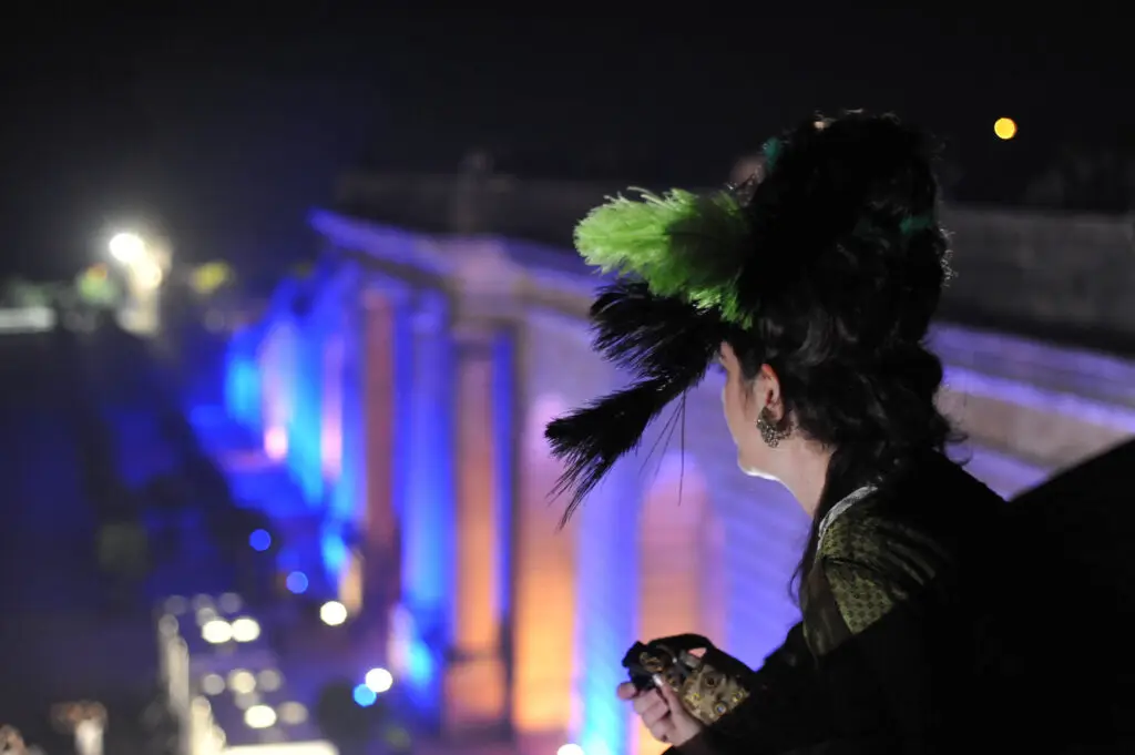 A woman in 18th-century costume with a feathered headpiece gazes over the illuminated grounds of the Palace of Versailles on the night of the masked ball of Versailles, holding a decorative mask in her hand.