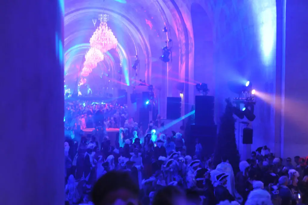 A grand crowd in period costumes and masks fills the Hall of Mirrors at the Versailles masked ball, illuminated by chandeliers and dramatic blue and purple stage lighting.