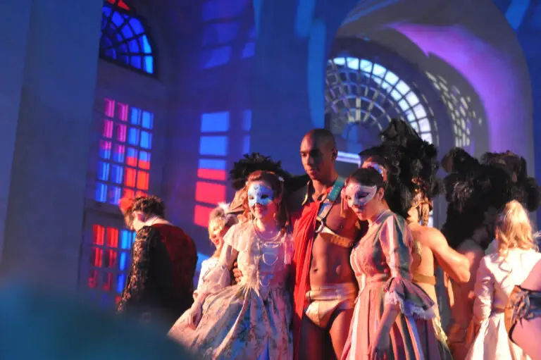 Masked guests in ornate 18th-century costumes pose with a muscular performer in gold briefs and a red sash under dramatic lighting at the Versailles masked ball, set against colorful reflections on arched palace windows.