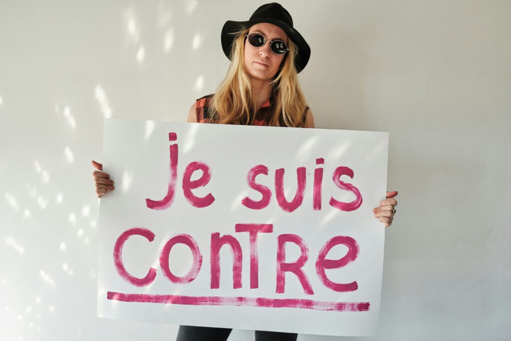 Person in dark sunglasses and a hat holding a bold sign that reads “Je suis contre” (“I am against”) — a modern portrait of the French art of complaining.