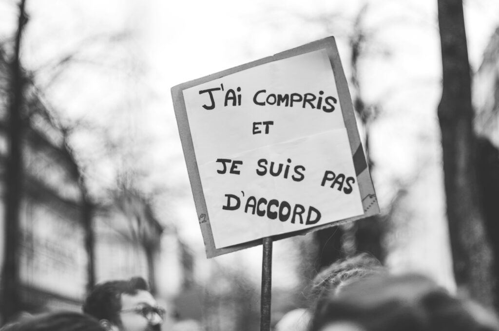 Protester holding a sign in French that reads "J’ai compris et je suis pas d’accord" ("I understand and I don’t agree") during a demonstration, with blurred figures and trees in the background.