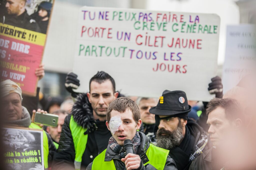 Yellow Vest protest in France with a man speaking into a microphone, his eye bandaged, and others holding colorful signs — a passionate display of civic resistance often dubbed the French art of complaining.