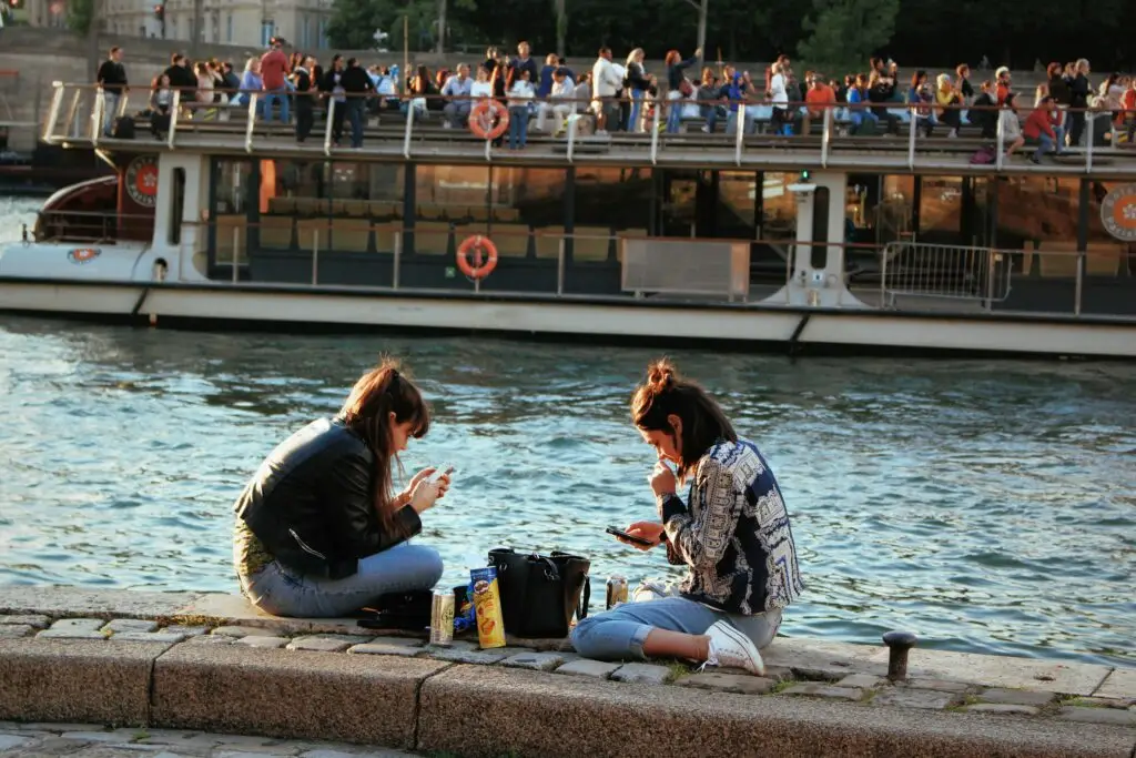 Two young women relaxing by the Seine River in Paris with snacks and phones, as a tourist boat passes by in the background.