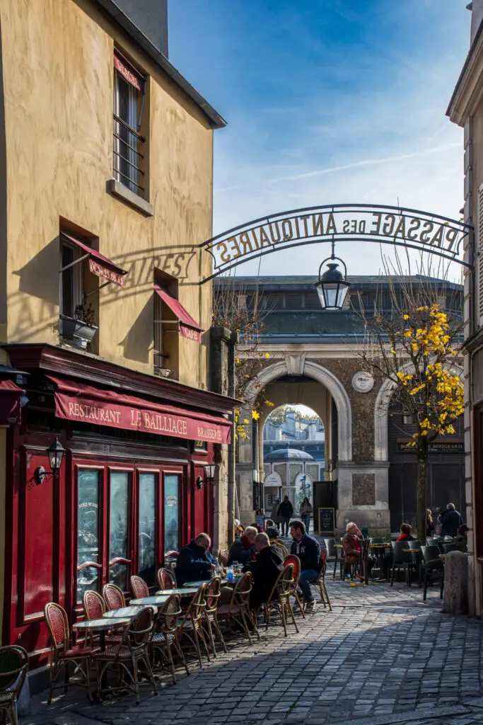 Outdoor café tables at Le Bailliage restaurant with people dining under the sign for Passage des Antiquaires in Versailles, France, a charming cobblestone alley known for antique shops and local shopping