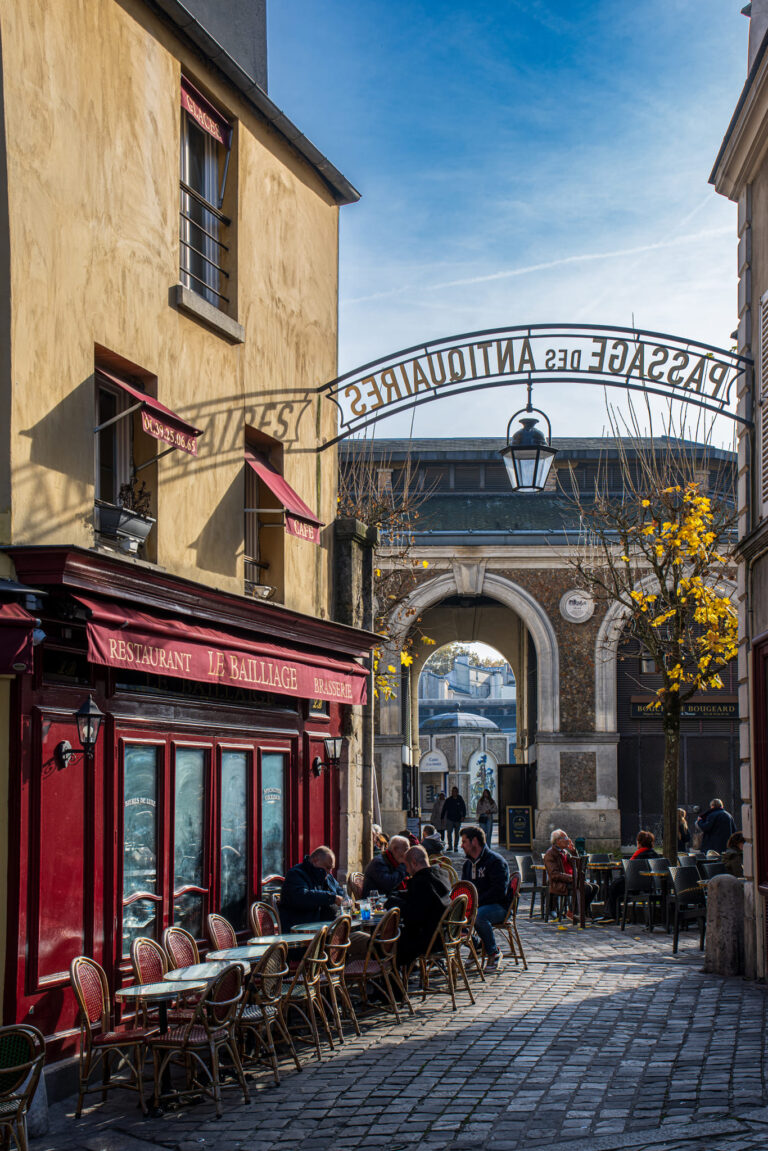 Outdoor café tables at Le Bailliage restaurant with people dining under the sign for Passage des Antiquaires in Versailles, France, a charming cobblestone alley known for antique shops and local shopping
