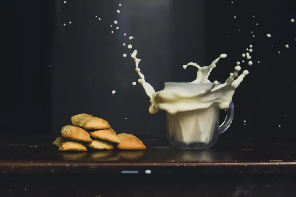 Glass of milk splashing dramatically beside a stack of golden French biscuits on a dark background.