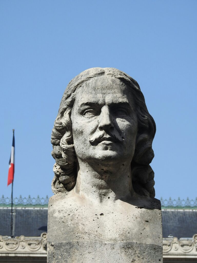 Stone bust of a stern-faced French man with flowing hair and a mustache, set against a clear blue sky with a French flag in the background — a face that looks like it disapproves of modern architecture.