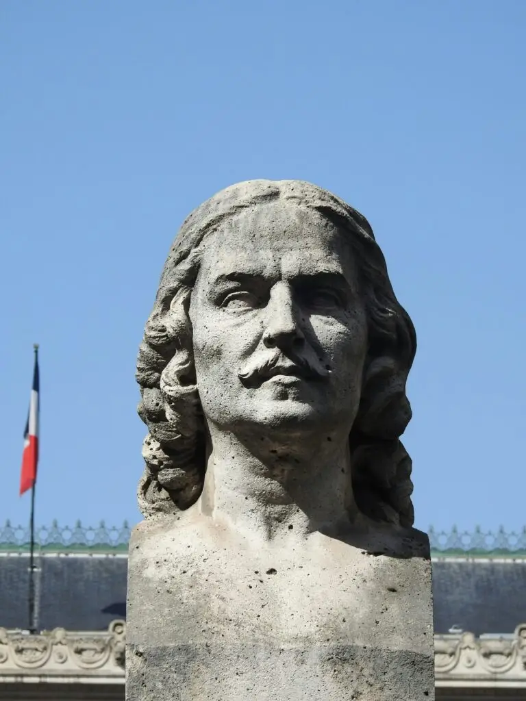 Stone bust of a stern-faced French man with flowing hair and a mustache, set against a clear blue sky with a French flag in the background — a face that looks like it disapproves of modern architecture.