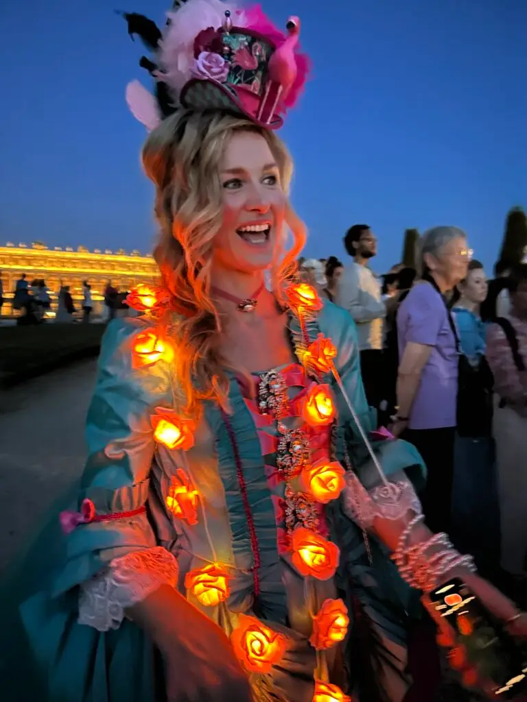 A joyful woman dressed in a vibrant 18th-century-style costume at the 2025 Masked Ball of Versailles. She wears a teal Rococo gown adorned with glowing orange rose lights and intricate lace cuffs. Her elaborate hat features pink feathers, flowers, and a miniature flamingo. The golden lights of the Château de Versailles glow in the background as a crowd of attendees, also in costume, mingles behind her during the twilight festivities.
