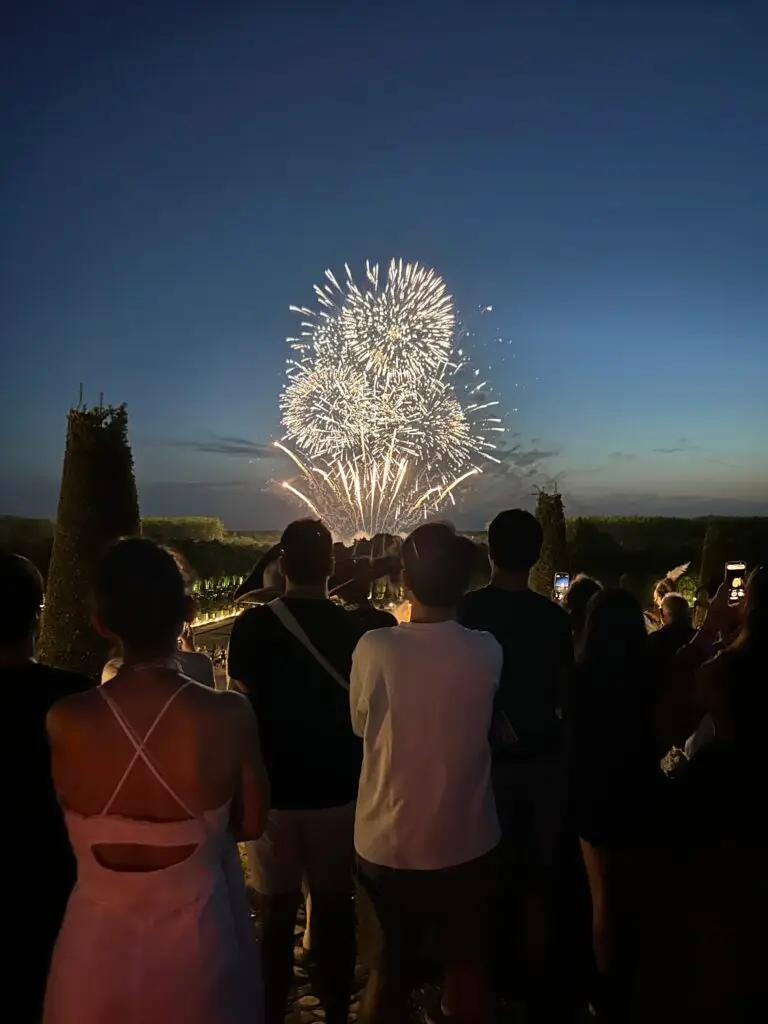 A crowd of spectators watches a dazzling fireworks display light up the twilight sky at the 2025 Masked Ball of Versailles. The bursts of golden sparks illuminate the horizon above the perfectly trimmed gardens of the Château. Silhouettes of guests, some in costume, stand in awe, capturing the moment on their phones as the grand finale unfolds against the fading blue of dusk.