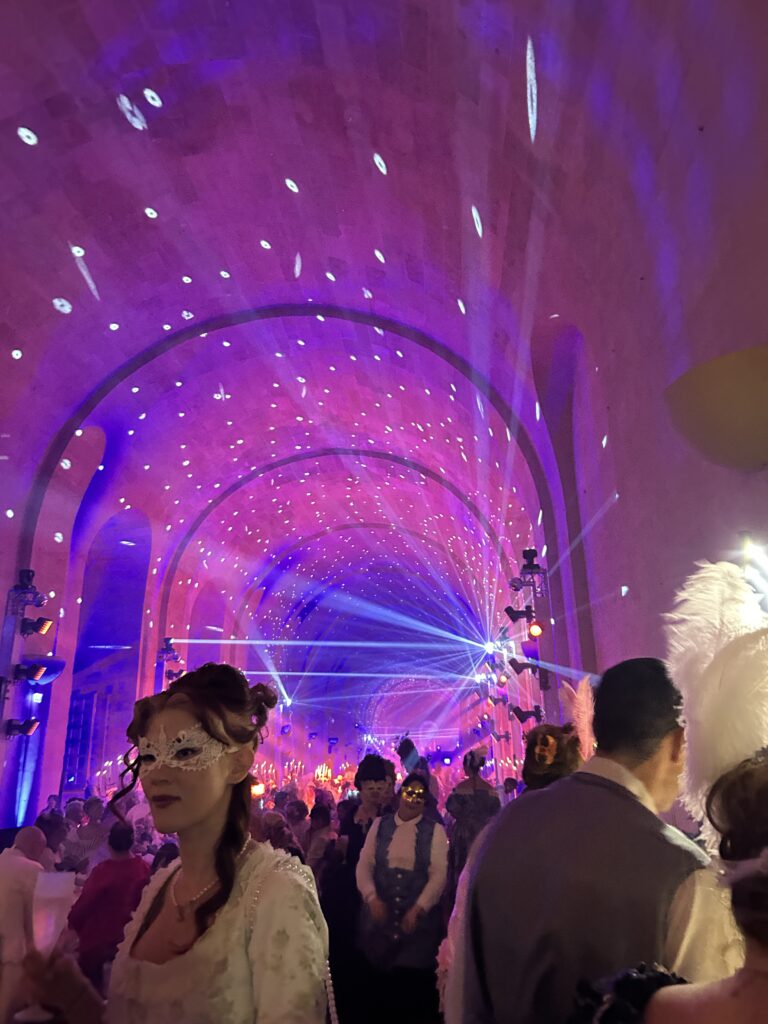 Guests in ornate 18th-century costumes and lace masks gather under a vaulted ceiling illuminated with soft pink, purple, and blue lights at the 2025 Masked Ball of Versailles. Laser beams and sparkling projections fill the air, creating a magical, starlit ambiance. Chandeliers and candlelight add to the opulence as attendees enjoy the blend of historical grandeur and modern spectacle in the heart of the palace.