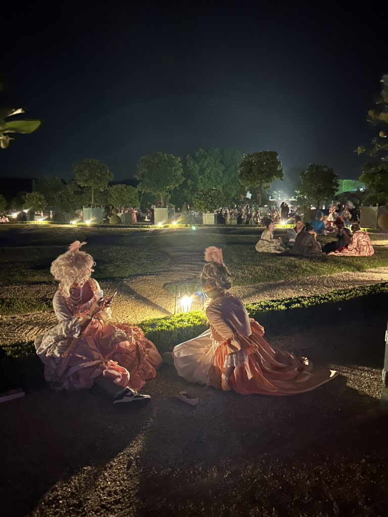 Two women in elaborate 18th-century gowns sit on the edge of a garden path at night during the 2025 Masked Ball of Versailles. Soft lantern light illuminates their pastel pink and peach dresses, adorned with lace, pearls, and bows. One guest checks her phone while the other gazes into the distance. In the background, more costumed attendees lounge on the manicured lawns beneath neatly trimmed trees, enjoying a peaceful moment amid the magical atmosphere.