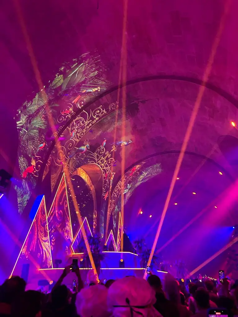 A vibrant stage scene at the 2025 Masked Ball of Versailles, filled with dazzling lights, neon jungle-themed projections, and swirling Baroque patterns cast onto the arched ceiling. Bright orange and pink laser beams crisscross the air as a crowd in powdered wigs and costumes watches the spectacle. The stage, decorated with palm trees and illuminated triangular structures, pulses with energy in a fusion of historical elegance and futuristic party vibes.