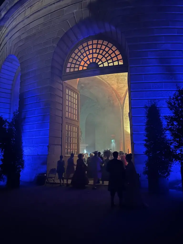 A grand arched doorway of a stone building at the Palace of Versailles glows with warm light from within, as costumed guests enter the 2025 Masked Ball. The exterior is bathed in dramatic blue lighting, creating a magical contrast. Silhouettes of attendees in period attire linger at the threshold, where Baroque elegance and modern theatrical lighting converge in a moment of enchantment.