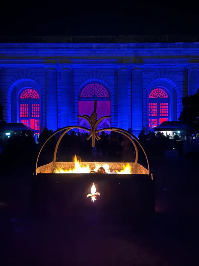A dramatic fire pit centerpiece featuring a glowing fleur-de-lis emblem burns brightly in the foreground at the 2025 Masked Ball of Versailles. Behind it, the majestic facade of a neoclassical building is illuminated in deep blue and pink lighting, with crowds of elegantly dressed guests gathering around. The scene blends regal symbolism, theatrical lighting, and the festive warmth of firelight to create a striking atmosphere under the night sky.