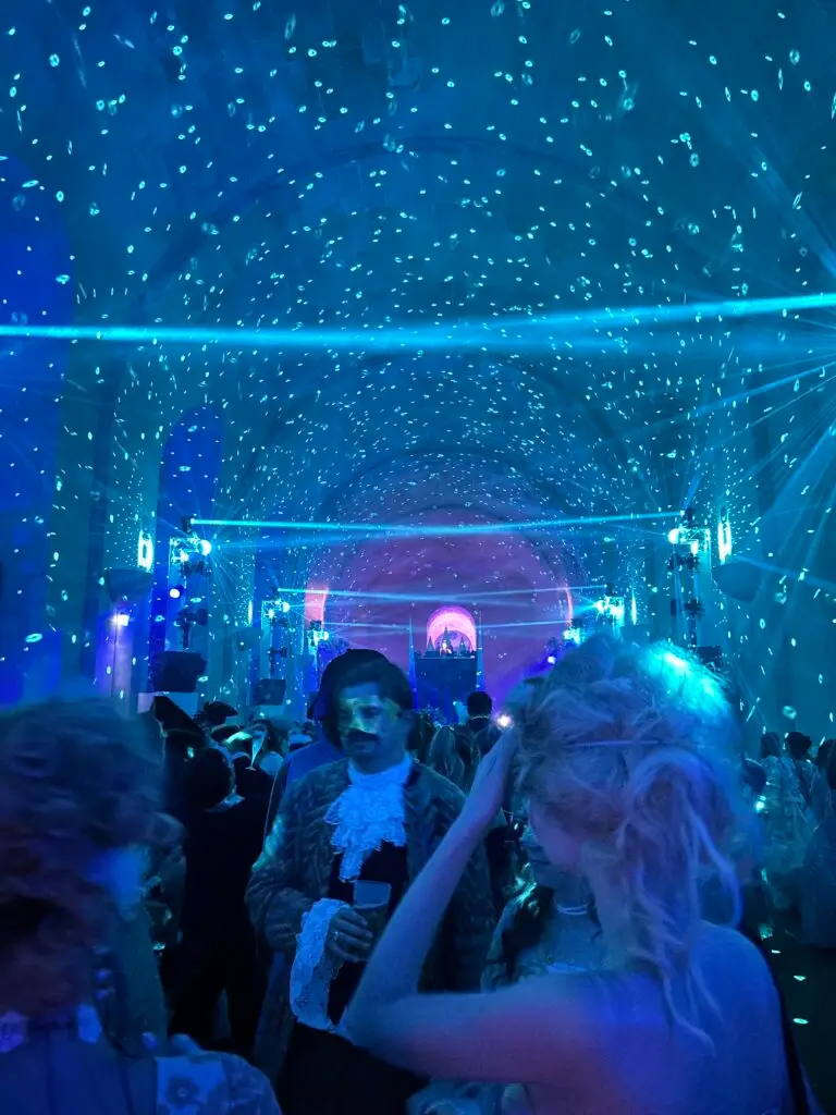 Costumed guests fill a vaulted hall bathed in blue and turquoise light at the 2025 Masked Ball of Versailles. Sparkling disco patterns dance across the ceiling, and laser beams crisscross above the crowd. A man in a baroque jacket and gold mask holds a drink while mingling with others dressed in powdered wigs and gowns. At the far end, a DJ booth glows beneath an arched alcove, blending historical opulence with modern nightlife energy.