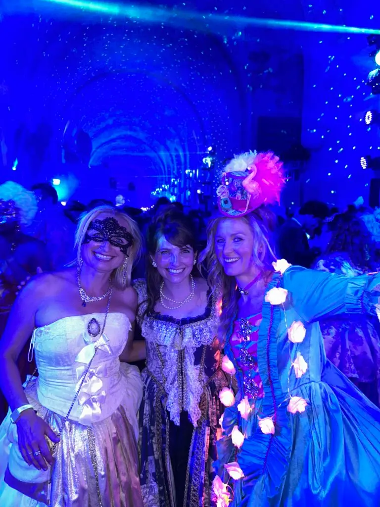 Three women in ornate Rococo-style gowns pose together under dazzling blue disco lights at the 2025 Masked Ball of Versailles. The woman on the left wears a strapless white and gold gown with a black lace mask and cameo necklace. The center woman dons a navy and cream lace dress with pearls, while the woman on the right is dressed in a teal gown with glowing rose lights and a flamboyant feathered hat. Behind them, the vaulted hall is filled with revelers, illuminated by starlike projections and laser lights.