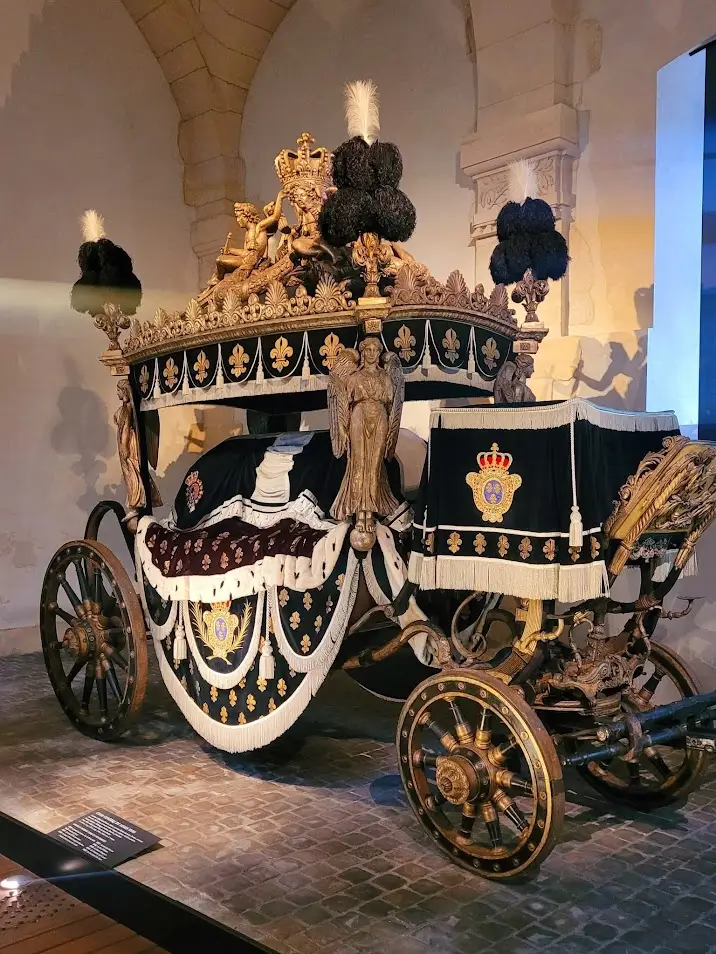 Majestic 19th-century royal funeral carriage displayed in the Coach Gallery at the Palace of Versailles, draped in black velvet with gold fleur-de-lis and crowned emblems, adorned with angel sculptures, feathered plumes, and elaborate bronze and gold detailing, set on a cobblestone floor.