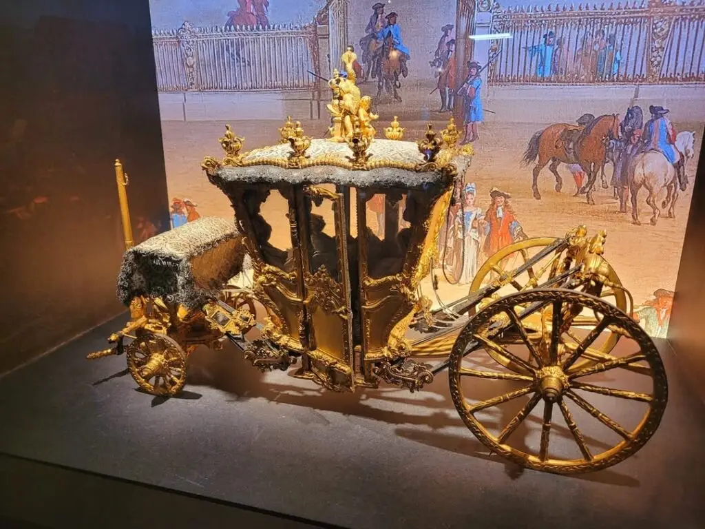 Ornate miniature royal carriage displayed in the Coach Gallery at the Palace of Versailles, featuring gold detailing, velvet trim, and a painted backdrop depicting 18th-century court life outside the royal stables.
