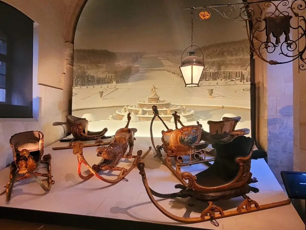 Display of elaborately carved and painted 18th-century royal sleds in the Coach Gallery at the Palace of Versailles, set against a winter scene of the Versailles gardens, featuring gilded animal figures, mythological designs, and luxurious seating for aristocratic use on snow-covered grounds.